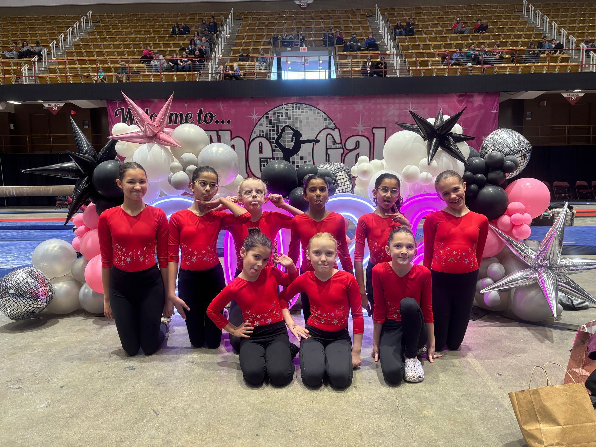 A group of gymnasts in red tops and black pants pose in front of a colorful balloon arch in an arena.
