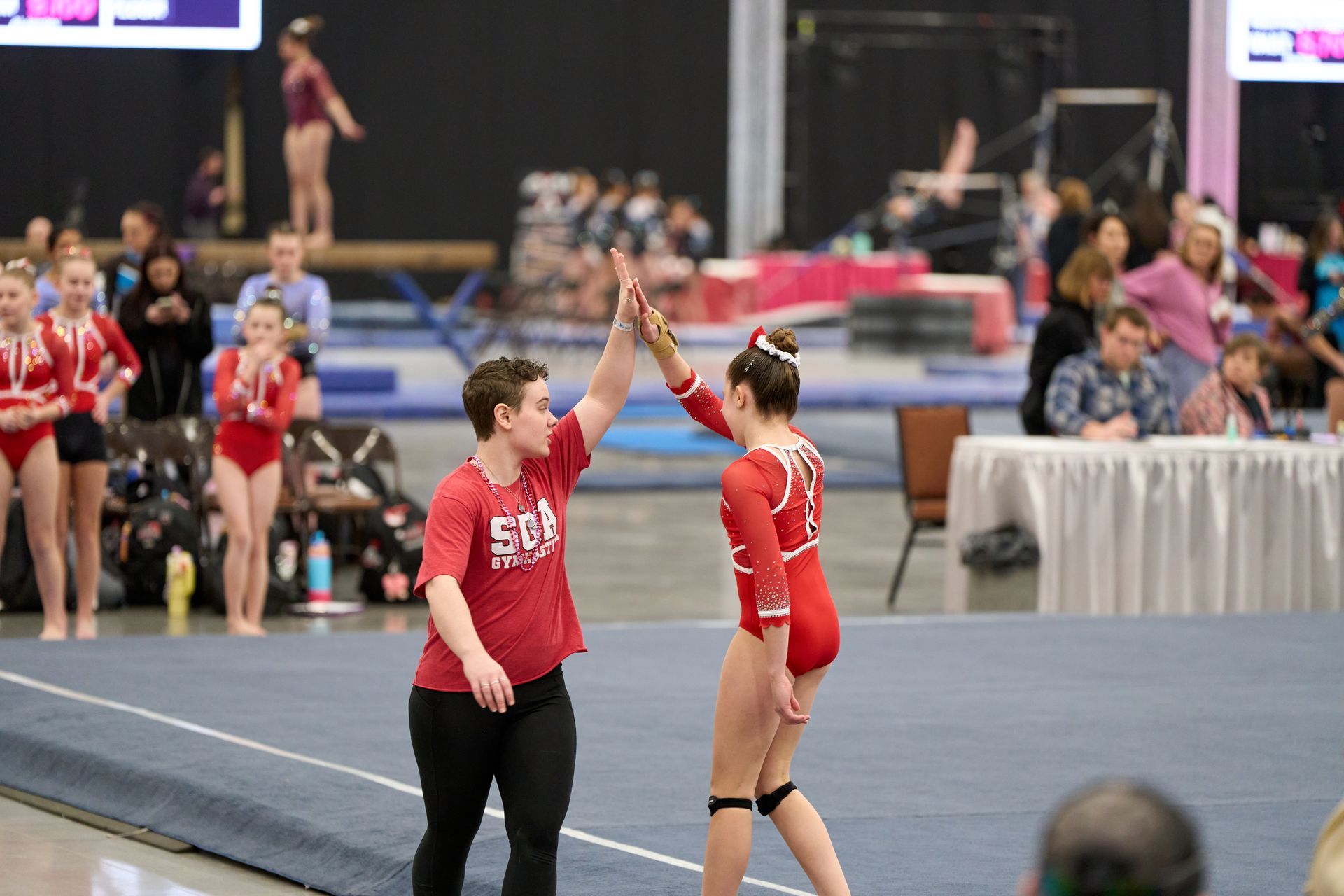 A gymnast in a red leotard and a coach in a red shirt high-five on a gymnastics floor during a competition.
