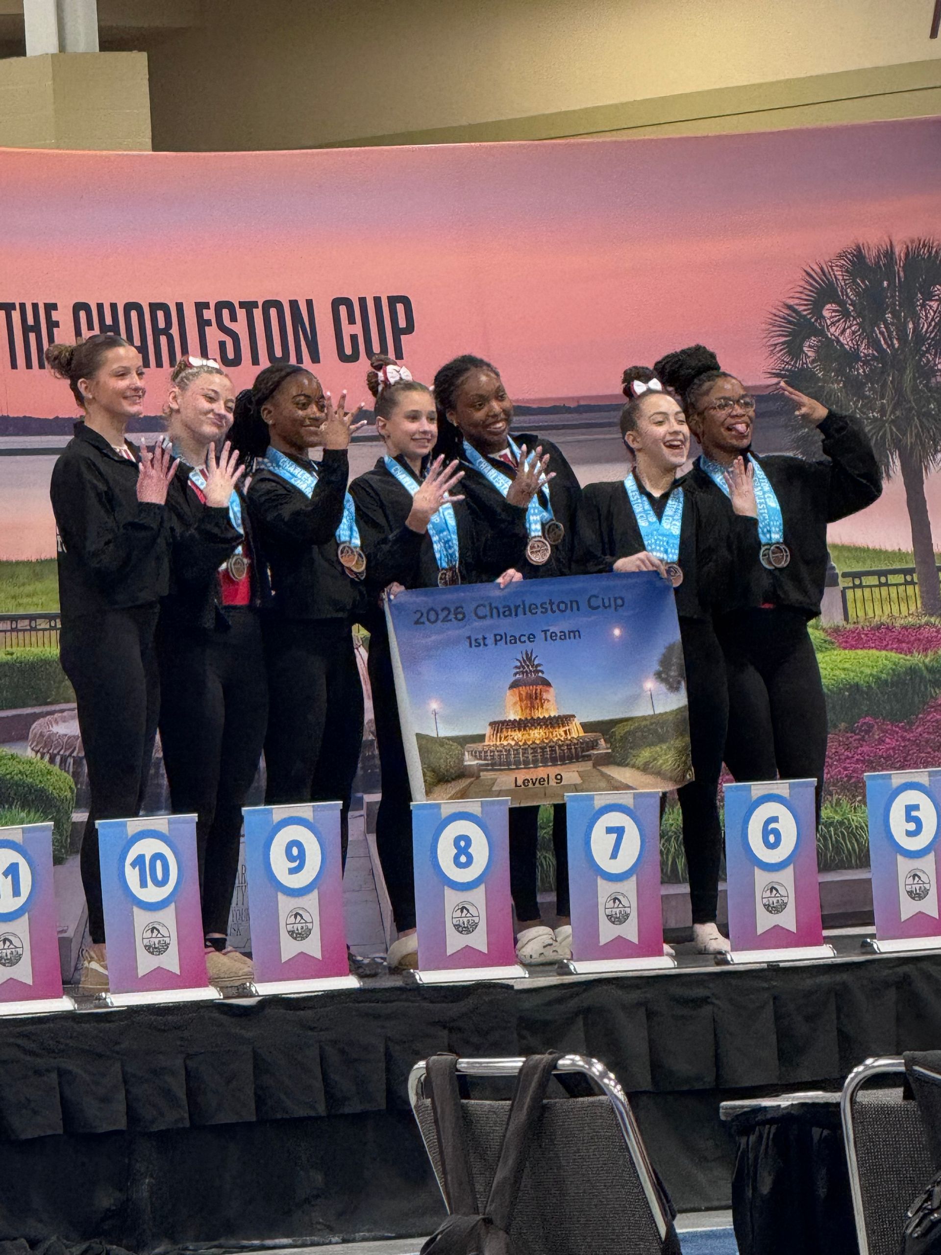 Seven gymnasts in black warmups stand on a podium labeled "The Charleston Cup," holding up a banner and smiling.