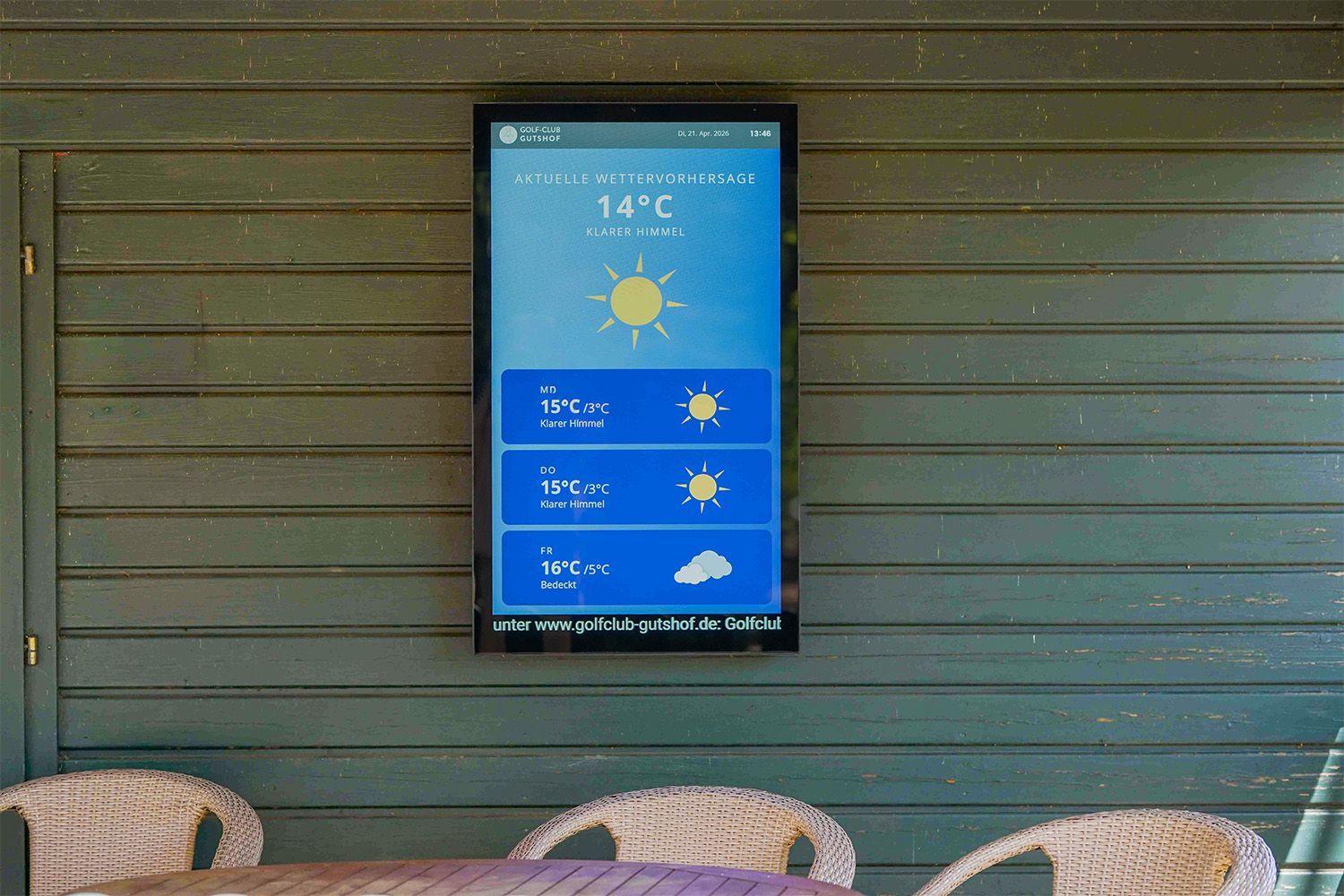 Digitale Wettervorhersageanzeige an einer Holzwand über Korbstühlen, die sonniges Wetter und 14 °C anzeigt.