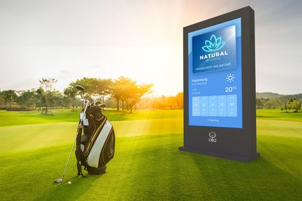 Auf einem grasbewachsenen Golfplatz steht neben einem digitalen Kiosk, der Wetterinformationen bei Sonnenuntergang anzeigt, eine Golftasche.