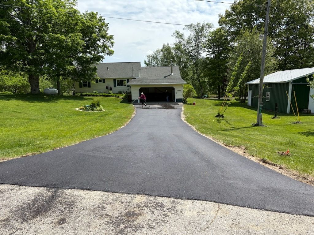 Black driveway leading to a house with an open garage, surrounded by green lawn and trees.