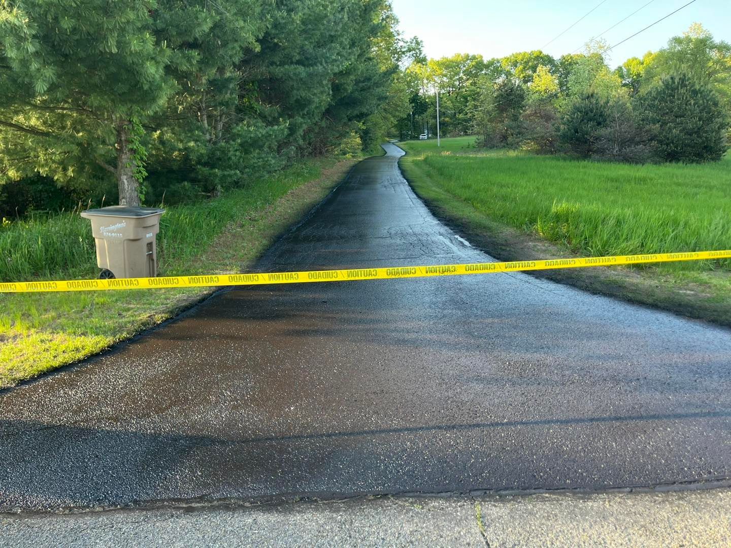 Yellow caution tape blocks a narrow paved road through a green rural area