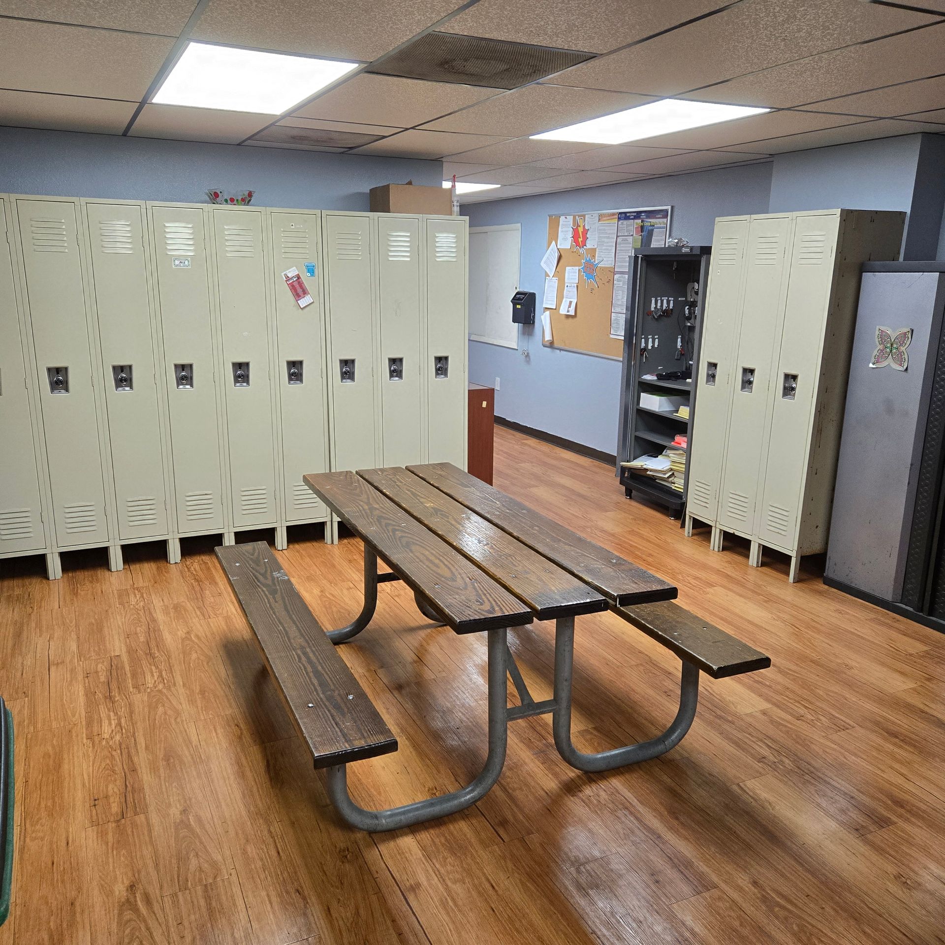 School locker room with beige lockers, wooden benches, and a picnic table on a wood floor.