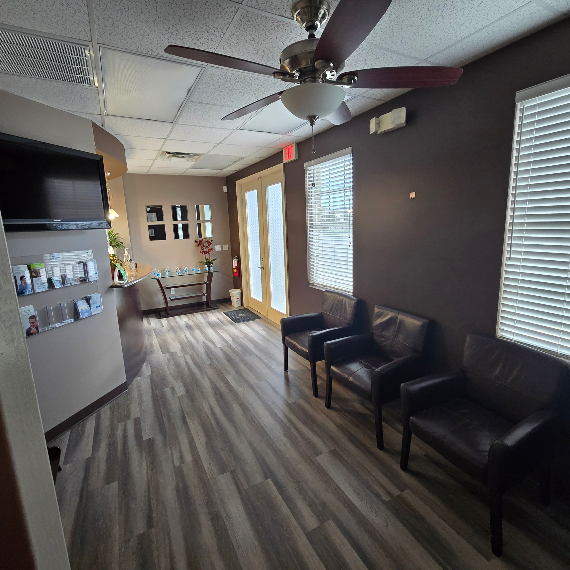 Waiting room with black chairs, ceiling fan, and windows with blinds along a gray wall.