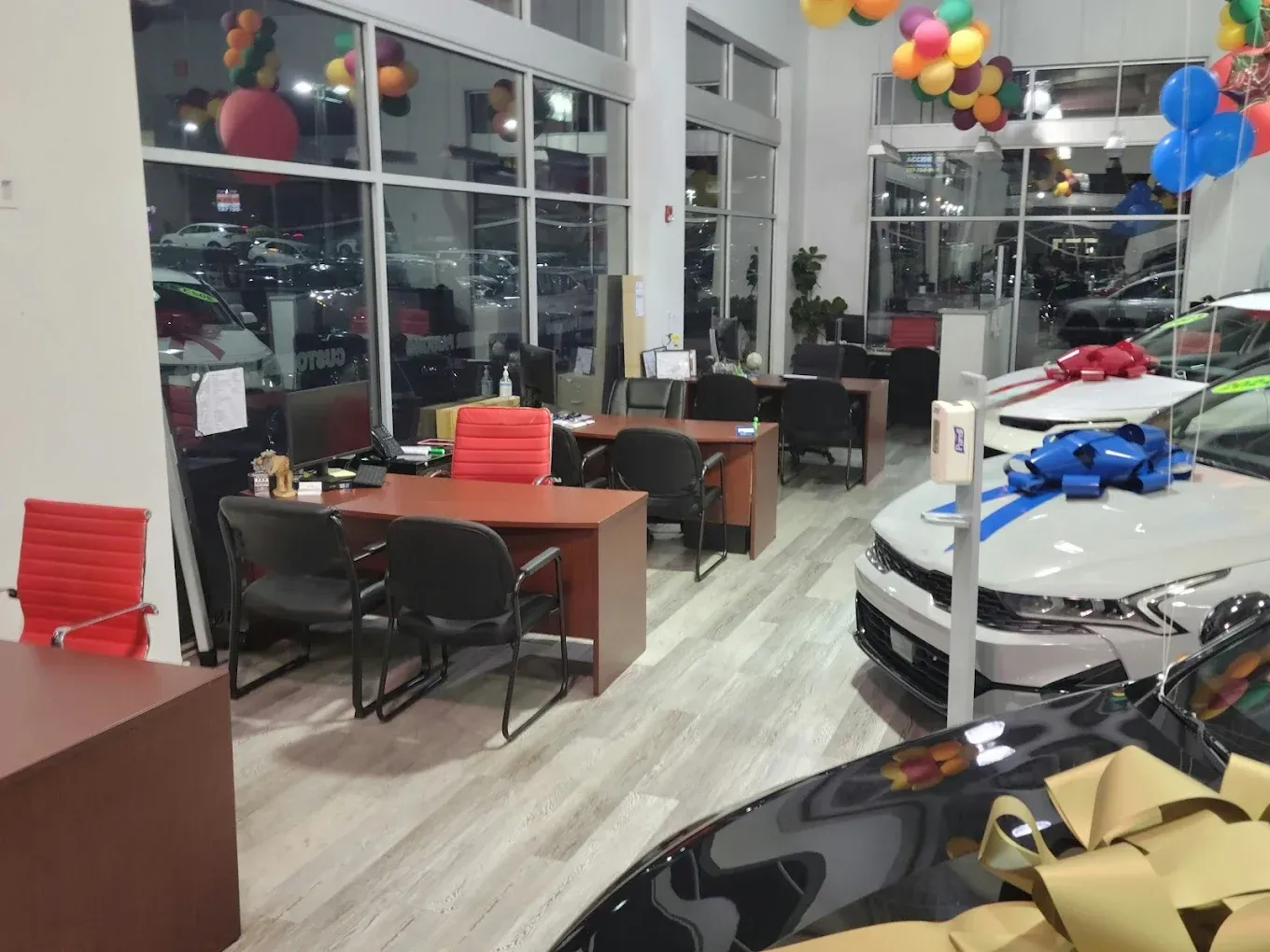 Inside a car dealership, desks with chairs, a car with a large bow, balloons, and glass windows.