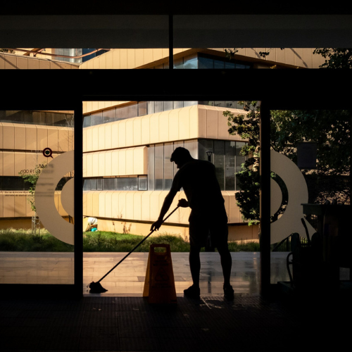 Silhouette of a person mopping inside a building entrance, warning cone present. Large windows and exterior building visible.