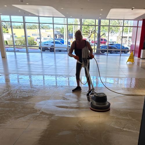 Person using a floor buffer to clean a large, light-colored tiled floor near large windows.