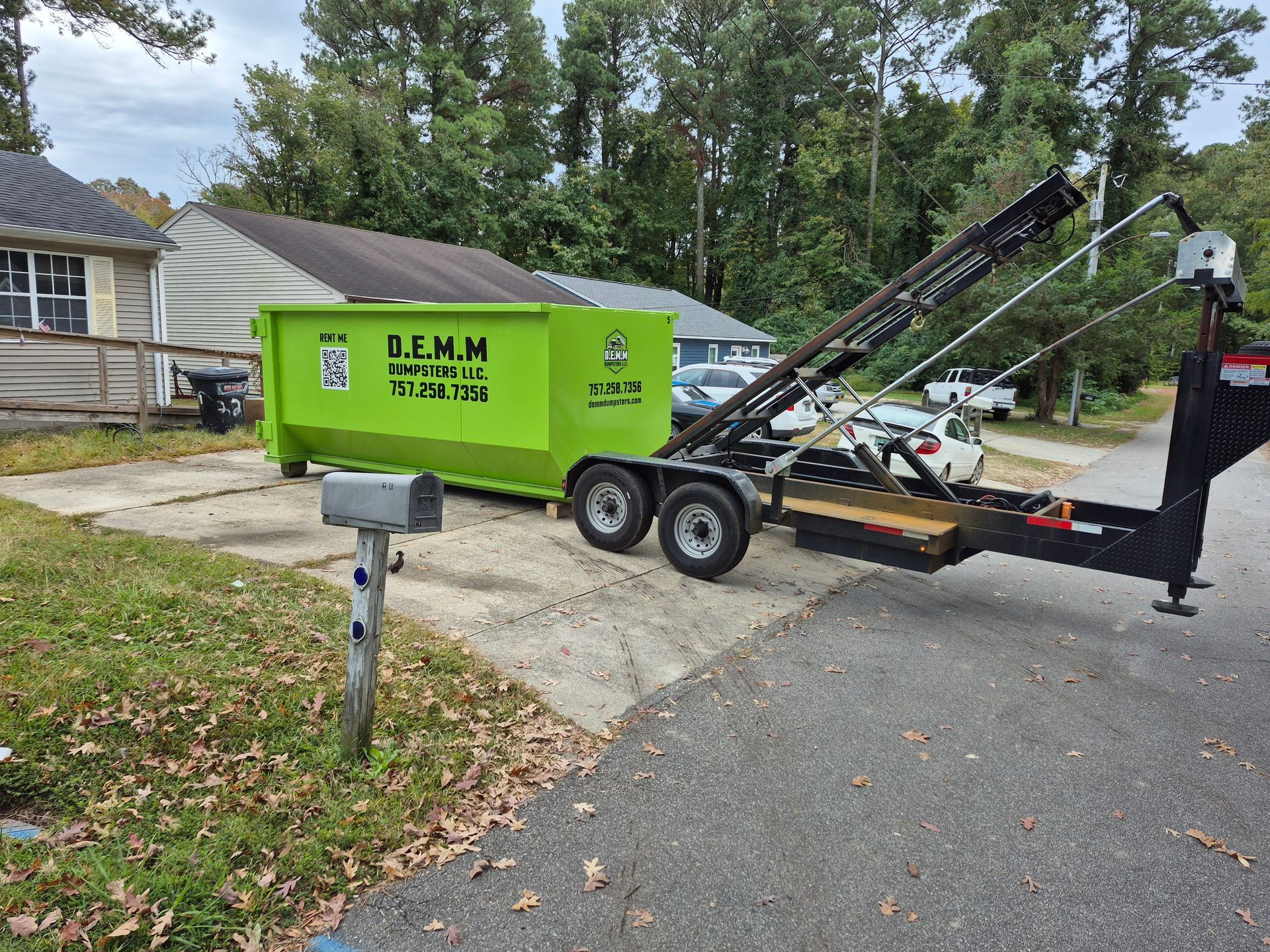 A blue pickup truck pulling a black dumpster trailer parked next to a house with a white garage.