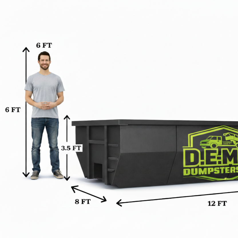 A blue pickup truck with a large black dumpster trailer parked in a driveway next to a brick house with a white garage door.