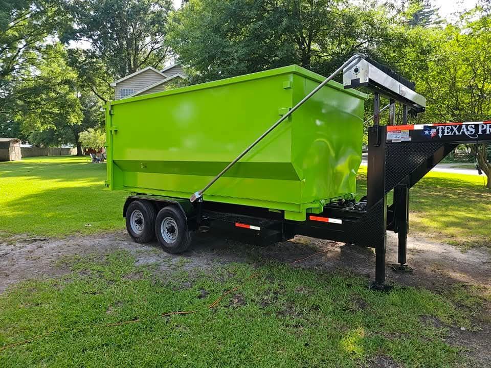 Green dumpster on a black trailer, parked on grass. Backyard setting with trees and a house.