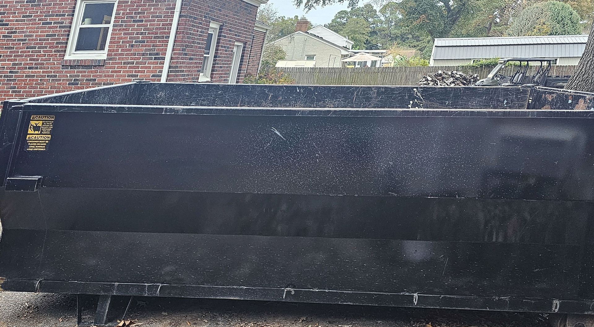 Black dumpster with overflowing debris sits outdoors in front of a brick building.