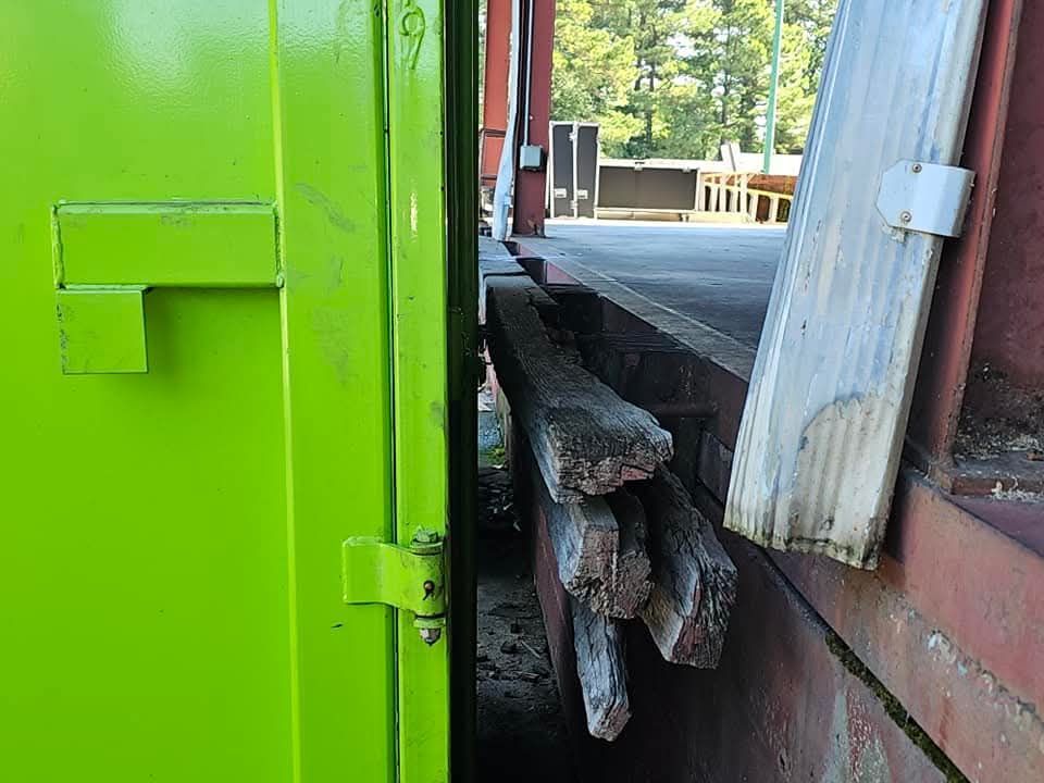 Green door next to a brick building with stacked, weathered wood. A piece of white wood leans against the building.