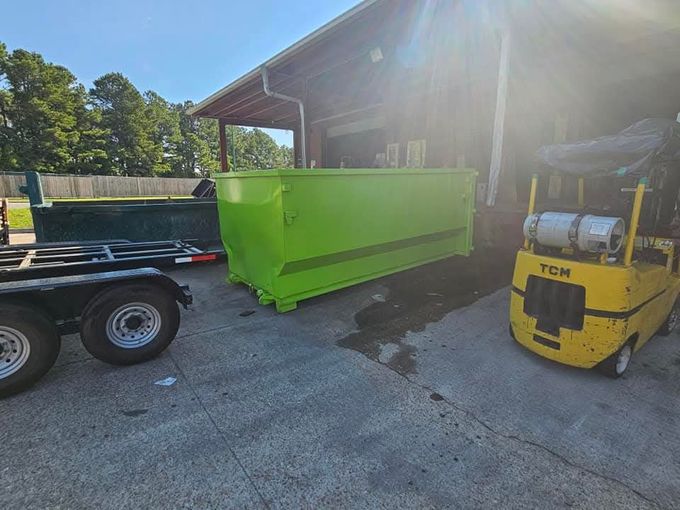 A bright green dumpster next to a yellow forklift and trailer in front of a building.