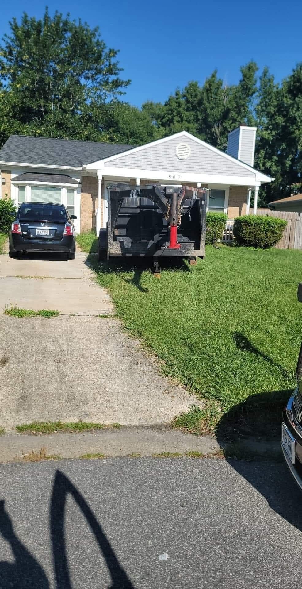 A black trailer parked on a driveway in front of a small, beige house. A car is parked to the left.