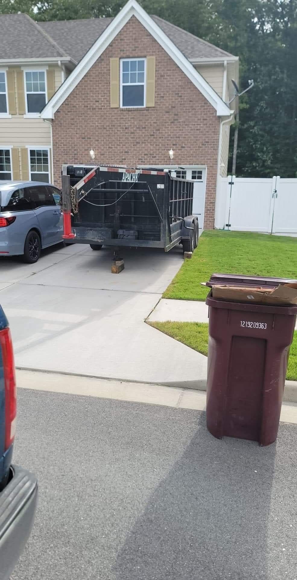 A dumpster trailer is parked in a driveway in front of a two-story house with a garage. A trash can is on the right.
