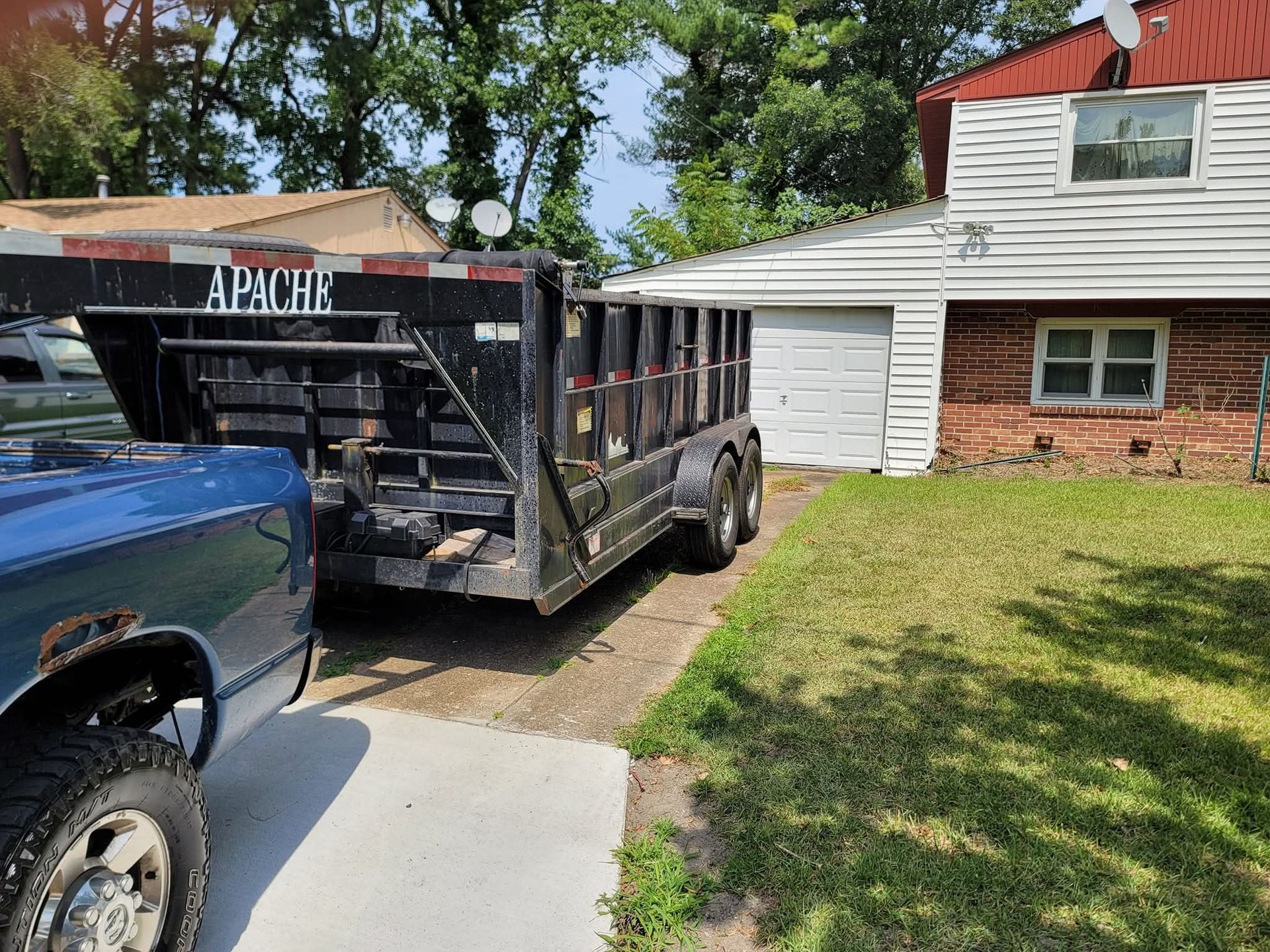 A blue pickup truck with a large black dumpster trailer parked in a driveway next to a brick house with a white garage door.