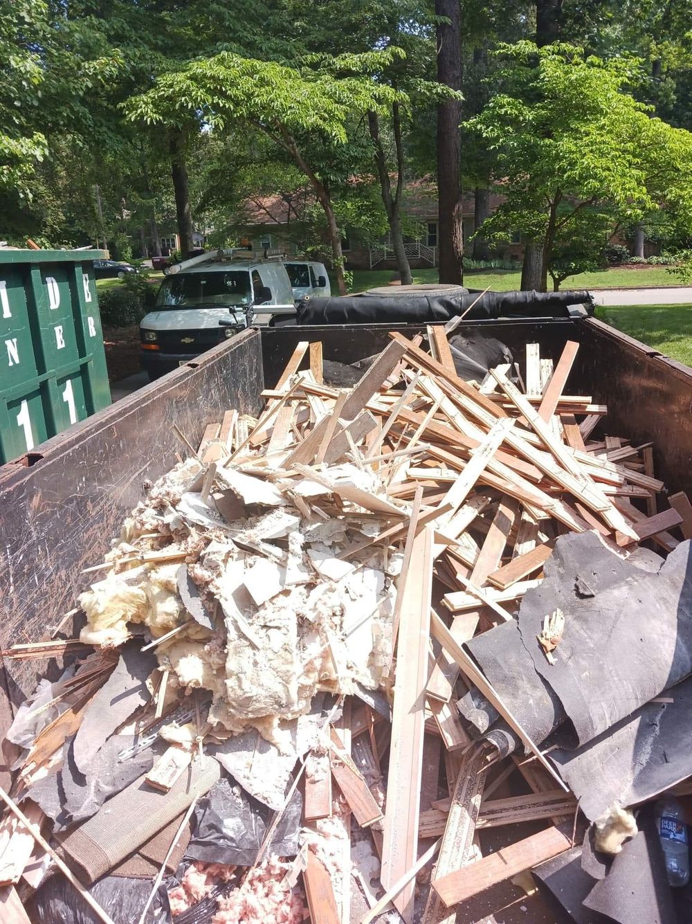 A dumpster filled with construction debris, including wood, shingles, and insulation, sits outdoors. A truck is visible in the background.