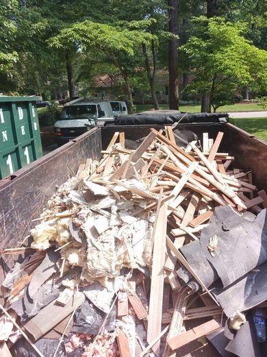 A dumpster filled with construction debris, including wood, shingles, and insulation, sits outdoors. A truck is visible in the background.