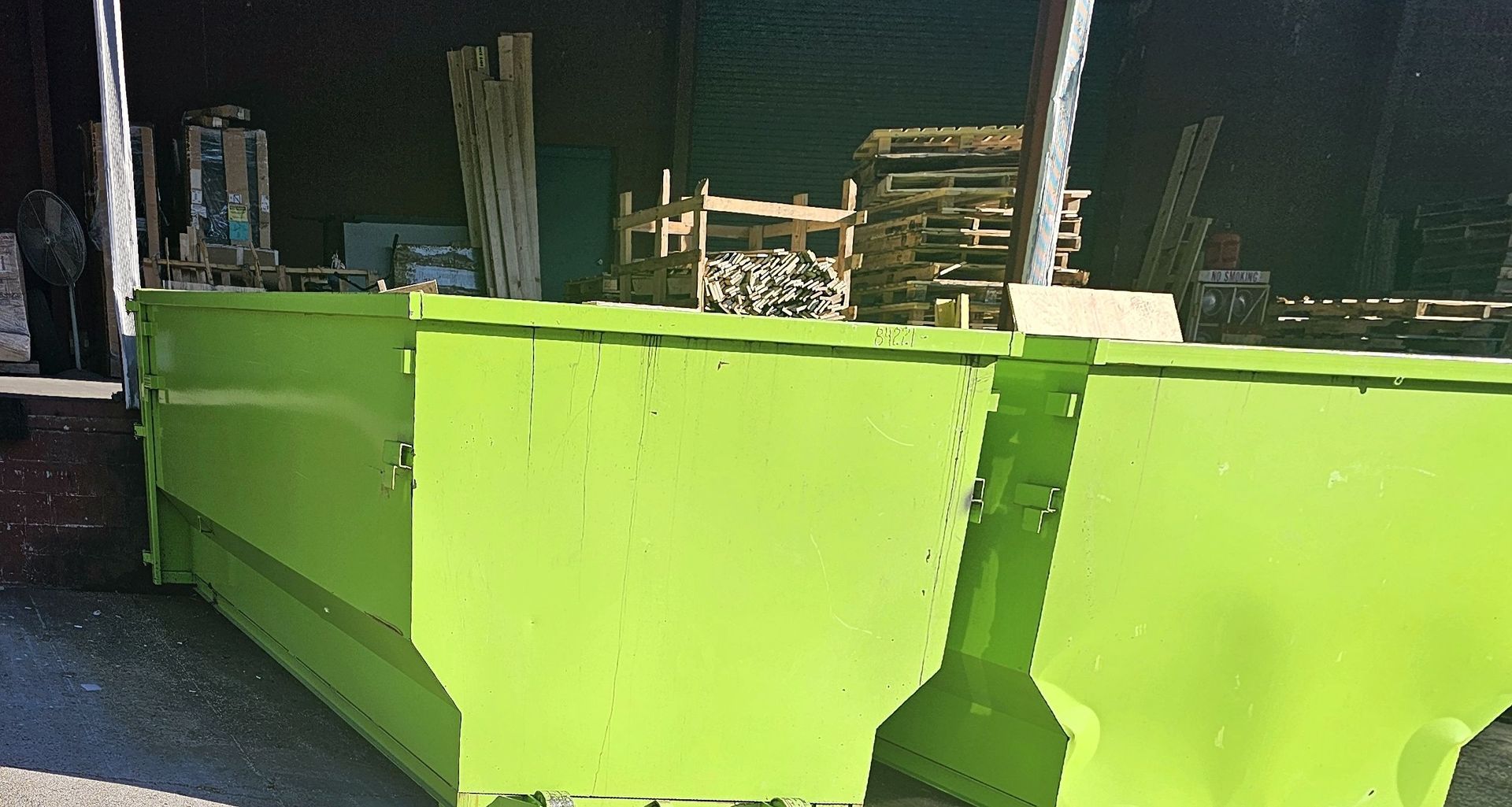 Two bright green industrial dumpsters sit outside, with lumber and pallets in the background.
