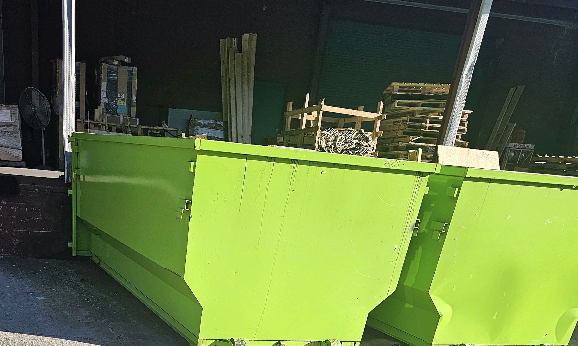 Bright green dumpster in an industrial setting, partially obscuring a cluttered background of wood and other materials.