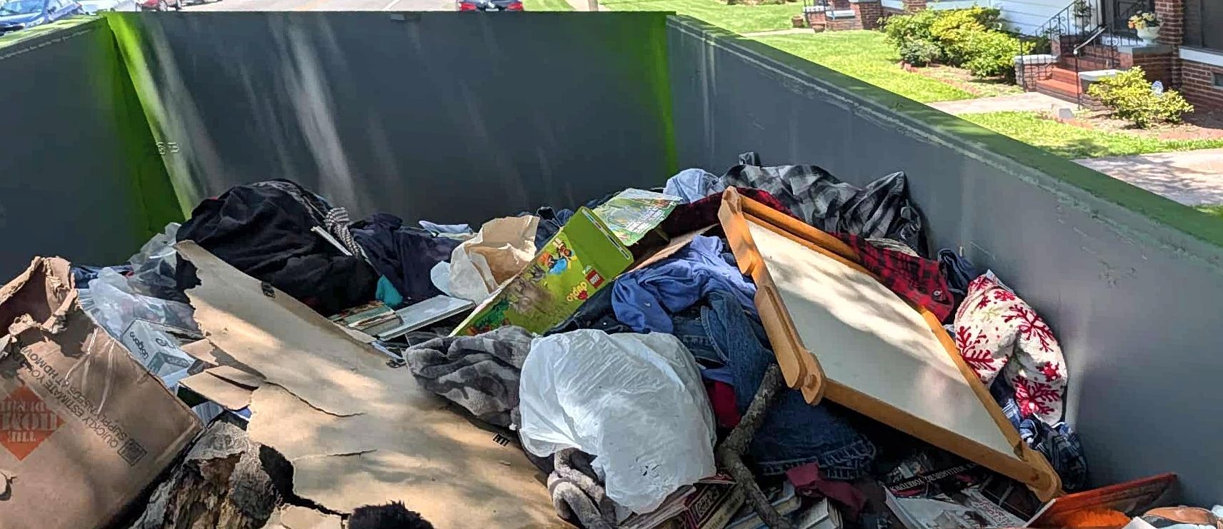 A dumpster filled with discarded items including clothing, cardboard, and a framed picture. Green walls surround the trash.