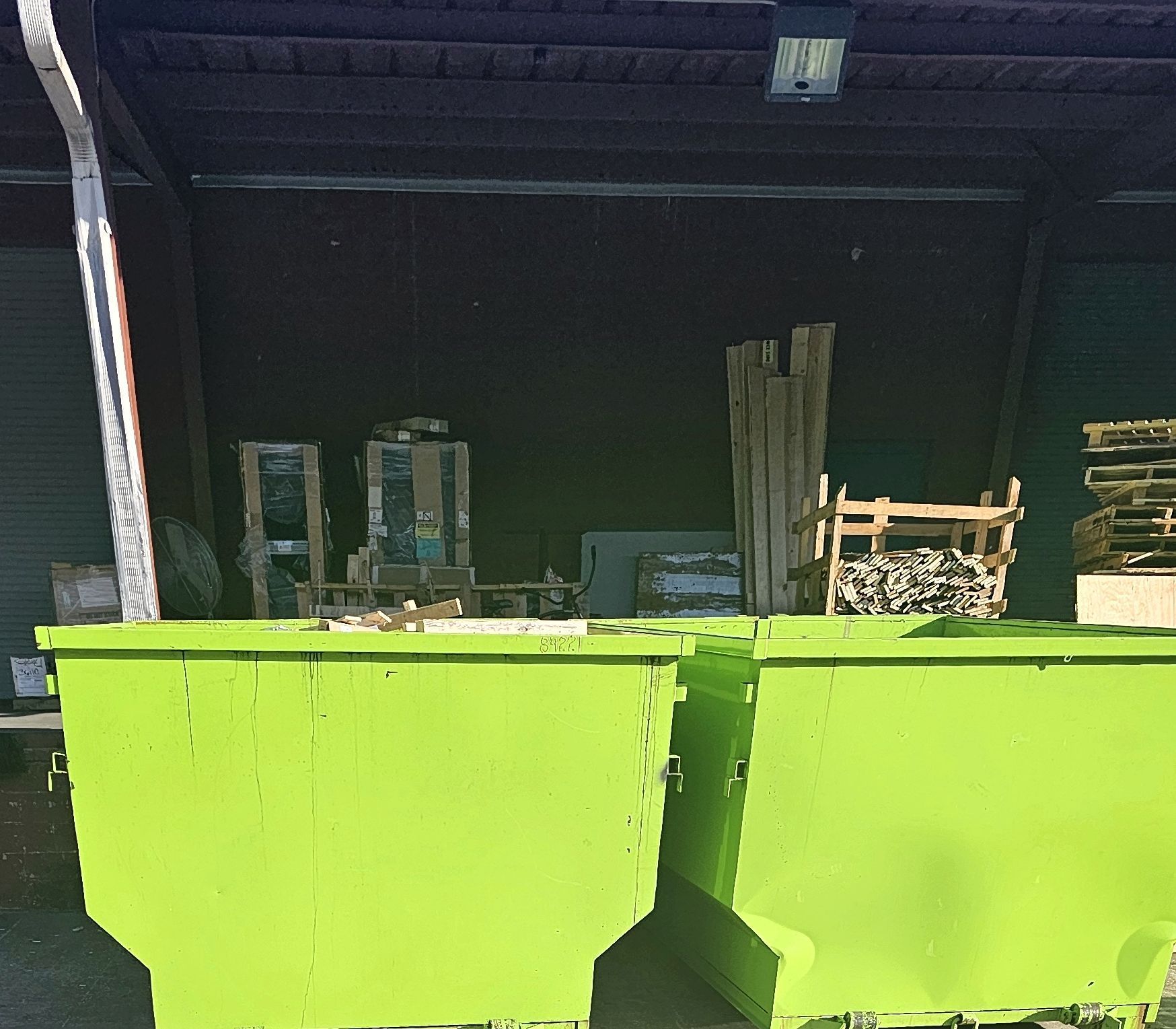 Two bright green dumpsters under a dark loading area, filled with wood and other construction debris.