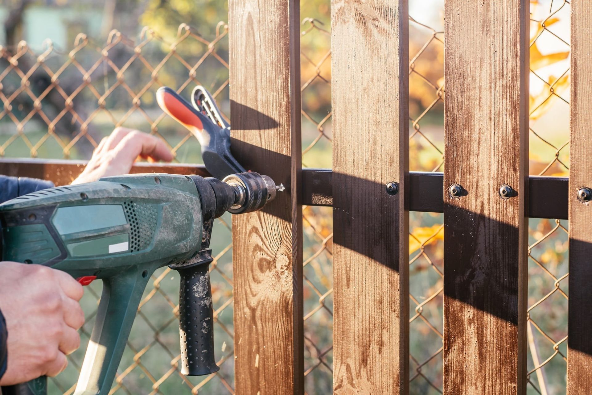 A person uses a green power drill to attach a wooden fence slat to a black metal horizontal support.