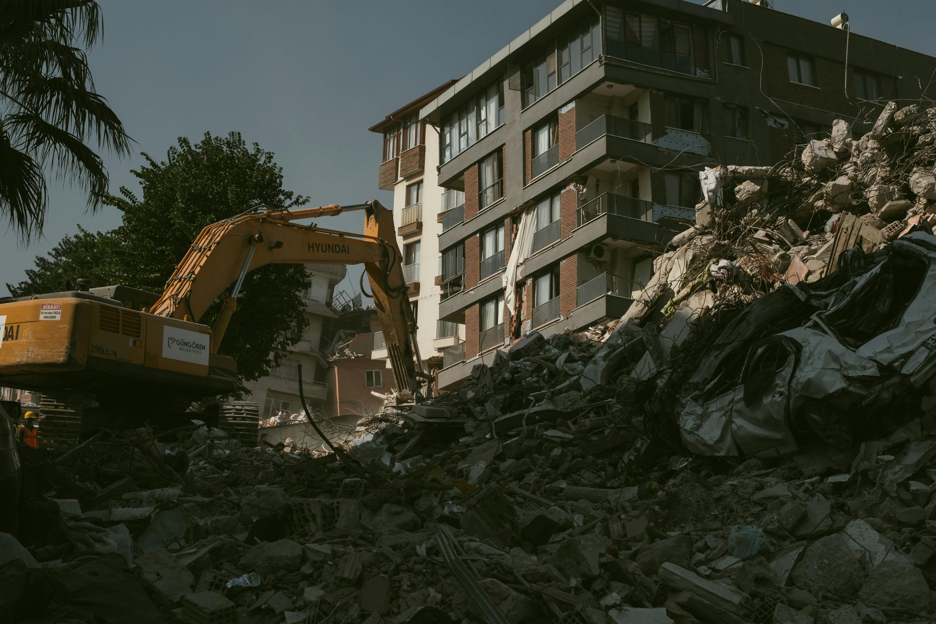 A yellow excavator clearing rubble and debris in front of a partially damaged modern building.