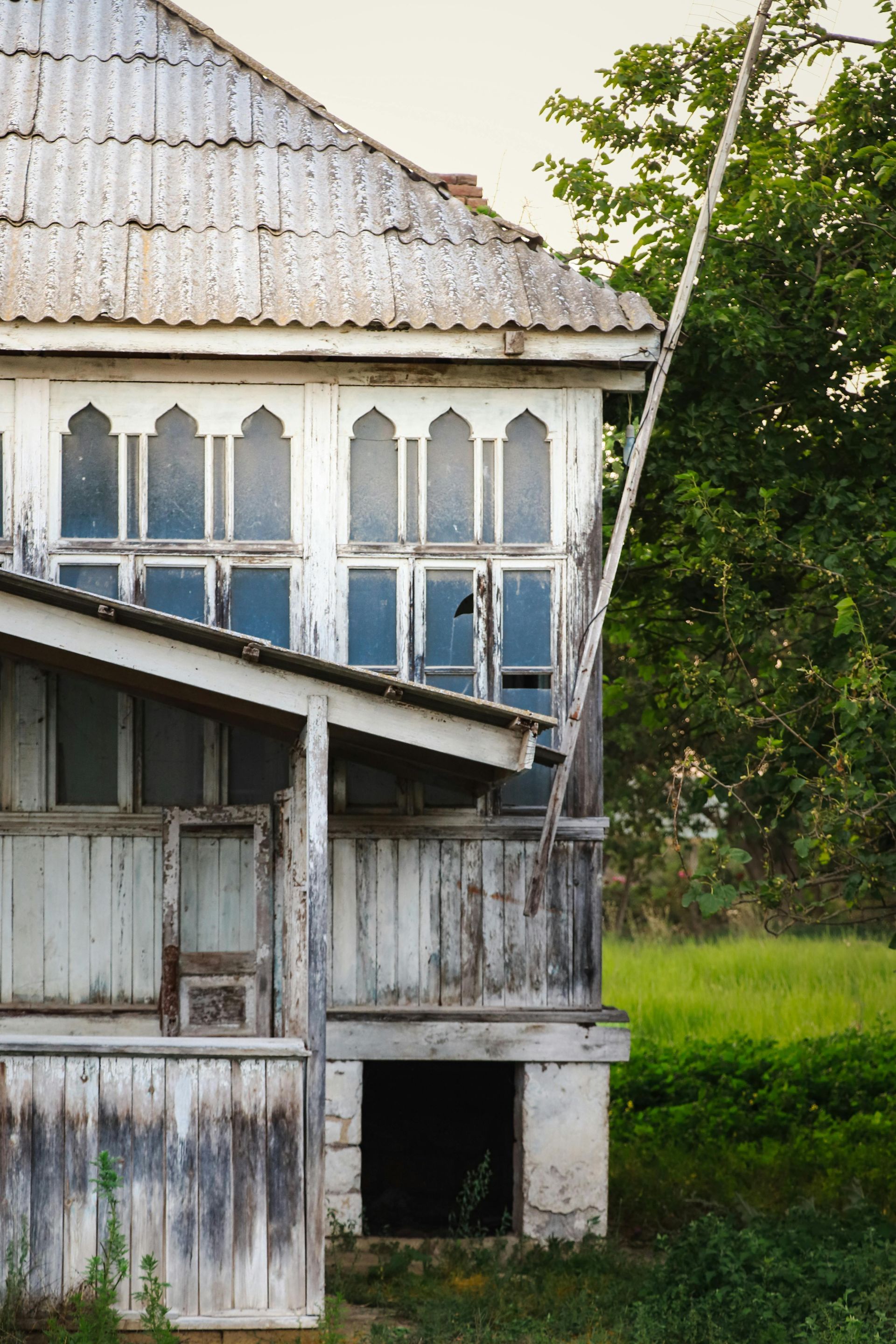 A weathered, white wooden house with a corrugated roof, ornate arched windows, and a shaded porch area.