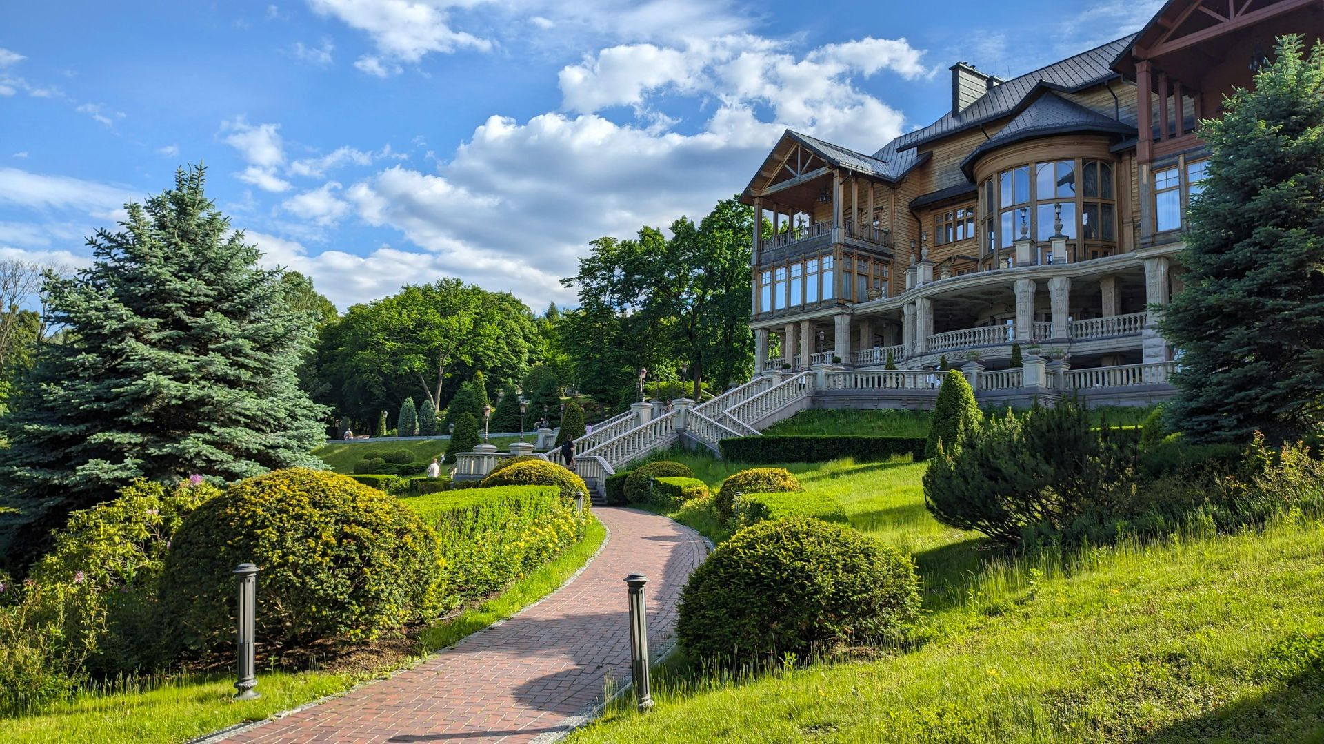 A grand, multi-story wooden mansion stands amidst lush green trees and manicured lawns, accessed by a stone garden path.