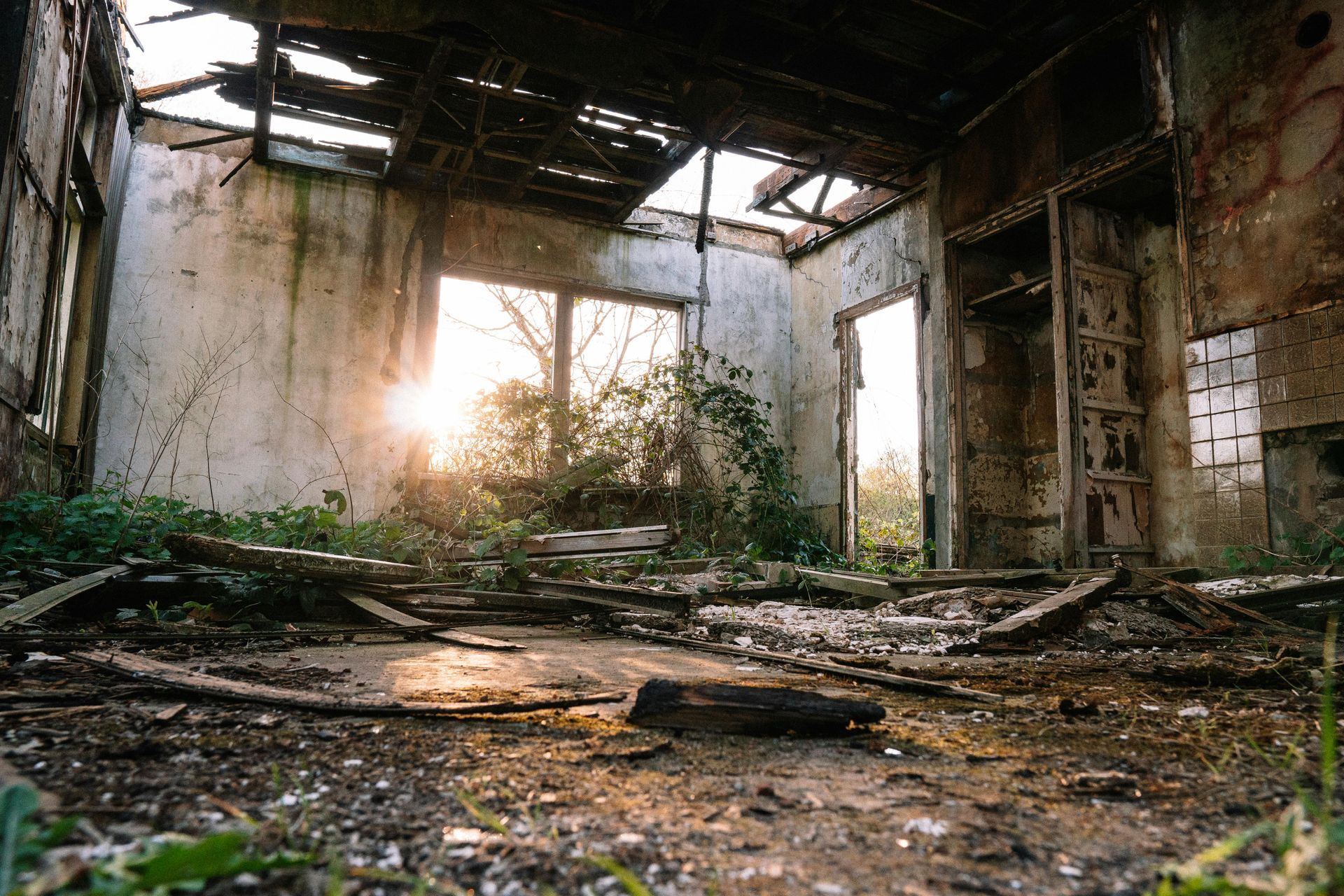 An abandoned, derelict room with a collapsed ceiling, debris-covered floor, and sunlight streaming through a large window.
