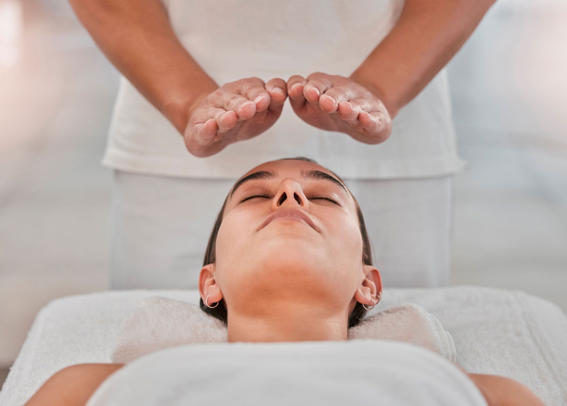 A person lies on a massage table with eyes closed, while hands are held hovering above their forehead in a healing gesture.