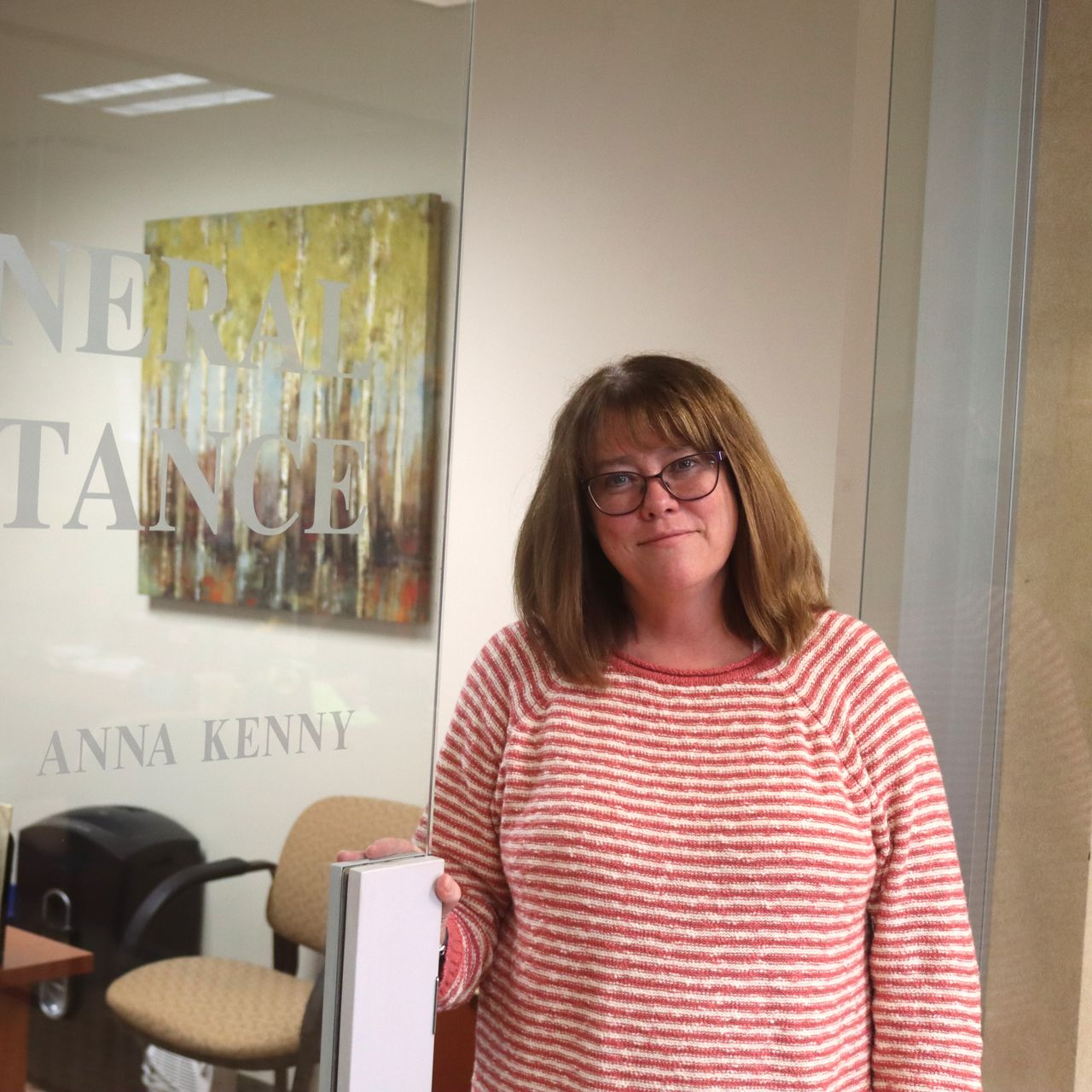 Woman in glasses, holding a white object, standing in front of a glass door with painting and text.