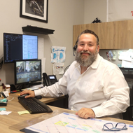 Man with a beard smiling at a desk in an office. Two monitors, keyboard, and phone are visible.