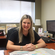 Woman seated at a desk, smiling with arms crossed; office setting with computer and papers.