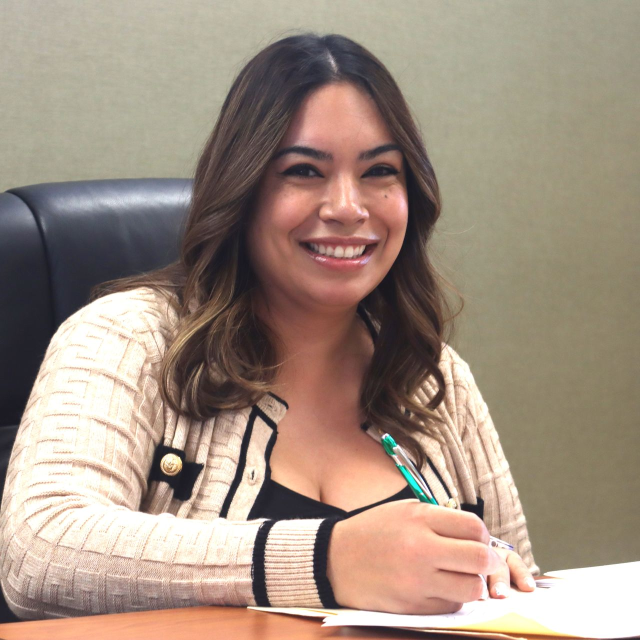 Woman smiling, writing at a desk, wearing a beige cardigan, black top, and holding a pen.