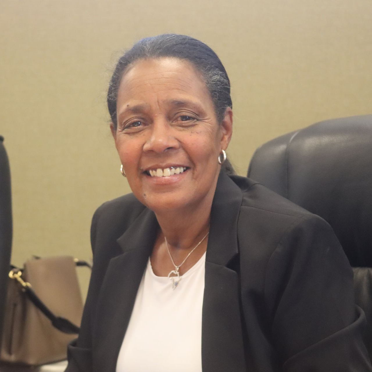 Woman smiling in a black blazer, white shirt, and silver jewelry. Seated near a purse.