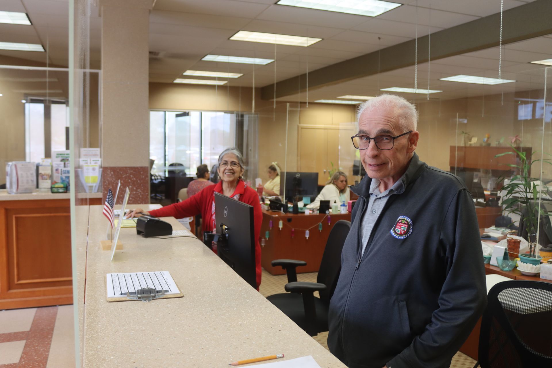 Two people at a counter in an office. A man in a blue jacket looks at the camera. A woman stands behind him.