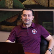 Man in a maroon polo shirt with laptop, sitting in an office. Artwork on the wall behind him.