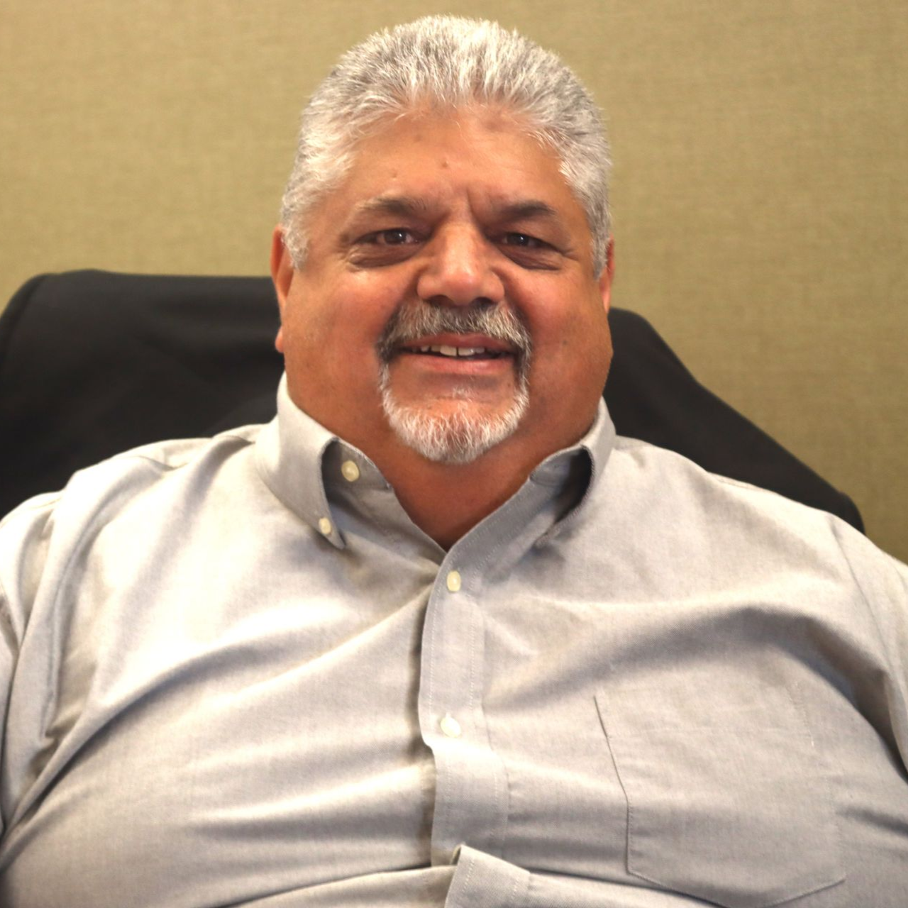 Man with gray hair and goatee smiling, wearing a light gray button-down shirt, indoors.