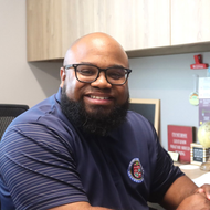 Man wearing glasses and a blue polo shirt smiles in an office setting.