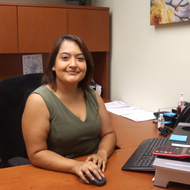 Woman in green top seated at desk, smiling. Office setting, computer and papers visible.