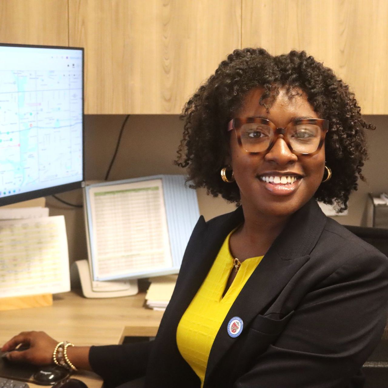 Woman smiling, wearing glasses and a black blazer, sitting at a desk with a computer.
