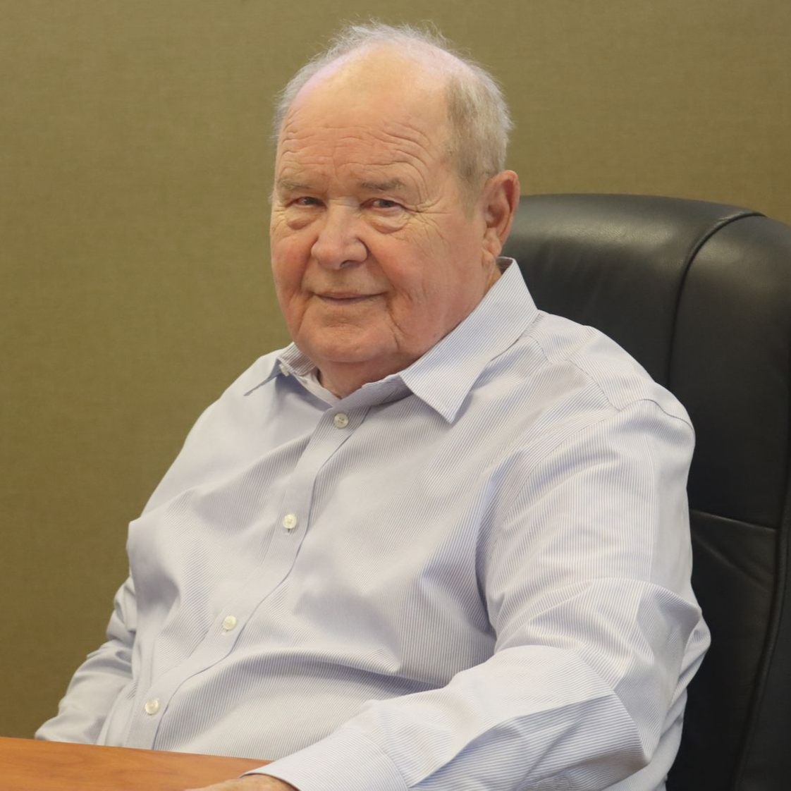 Man in light blue shirt seated in a black chair, smiling. Neutral background.