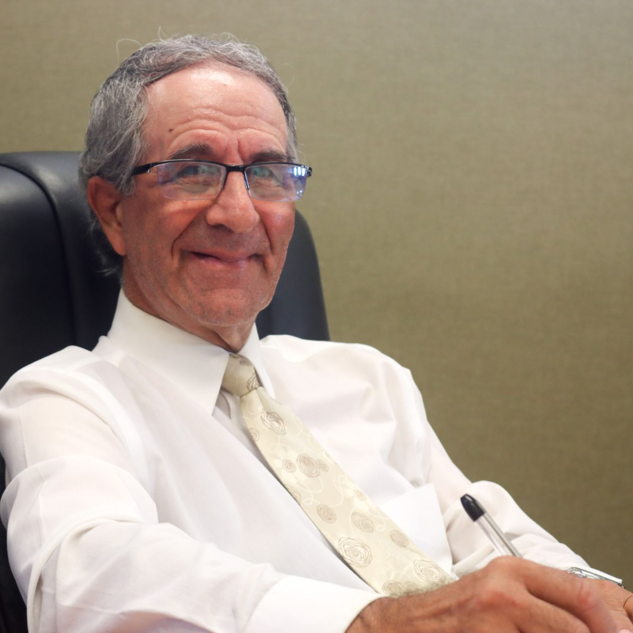 Man in glasses, white shirt, and tie, sitting in a chair and smiling.