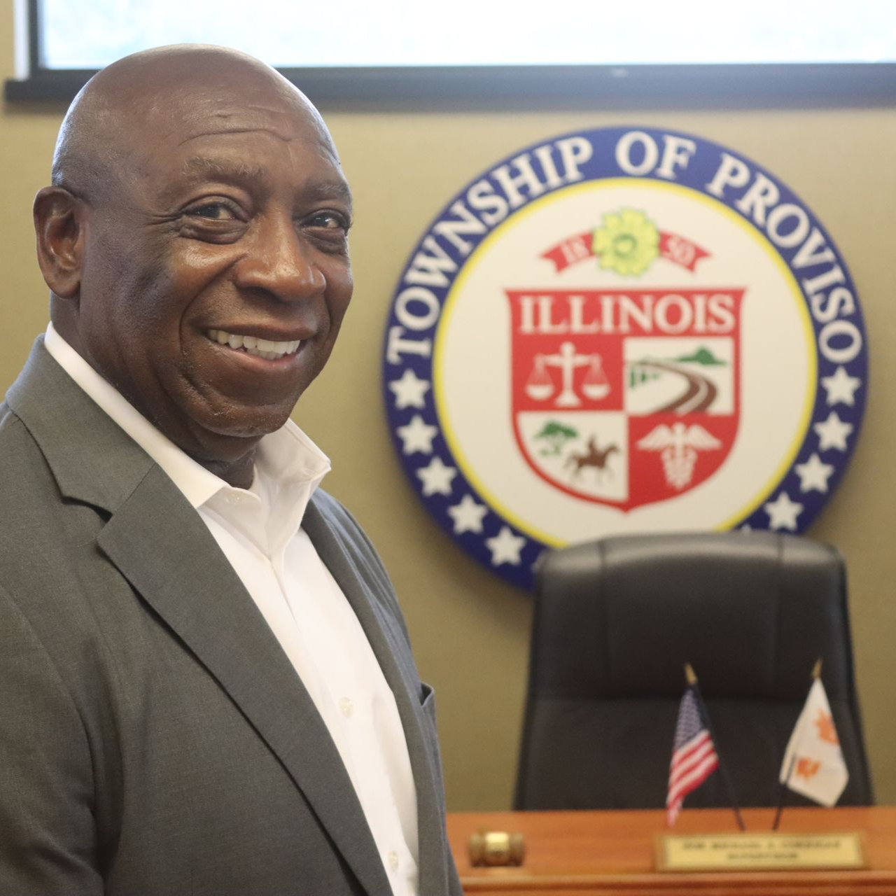 Man in grey suit smiles in front of a Township of Proviso, Illinois seal.