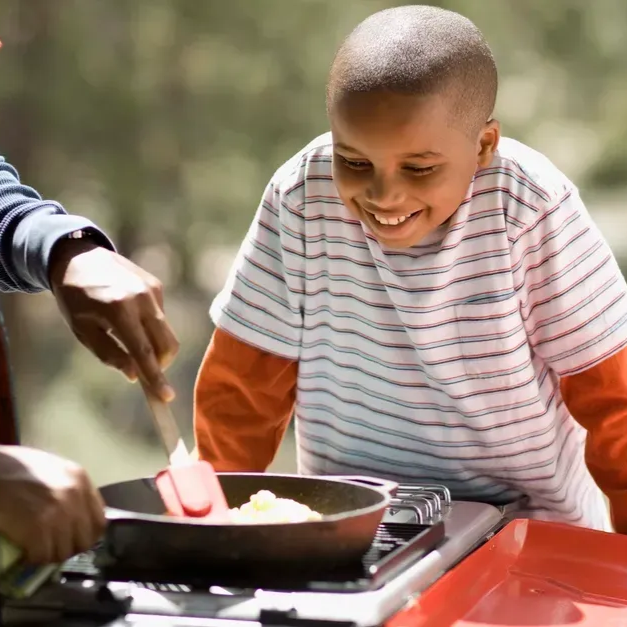 Boy watches as someone cooks in a pan outdoors. The person uses a spatula.