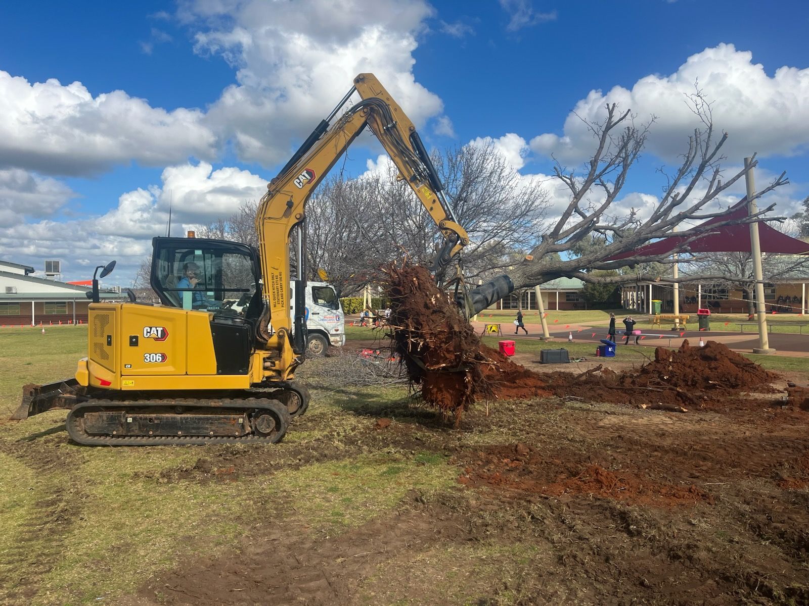 Yellow excavator removing a tree from a grassy area near a playground — Dubbo Premier Landscapes in Dubbo, NSW