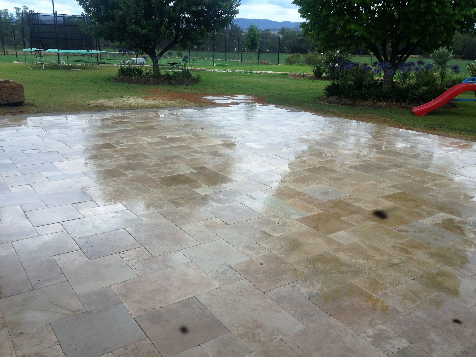 Wet stone patio reflecting a green lawn, trees, and cloudy sky — Dubbo Premier Landscapes in Dubbo, NSW